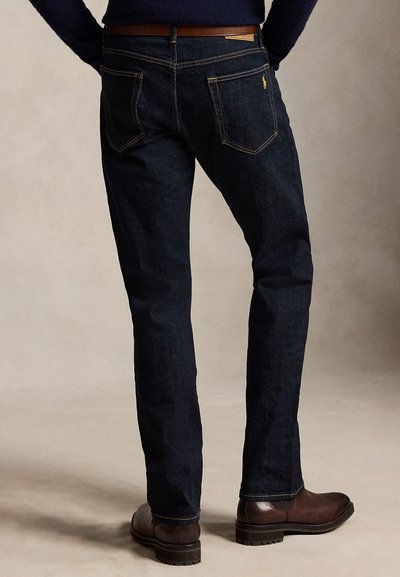 Zadný pohľad na osobu, ktorá má na sebe tmavomodré džínsy, hnedý opasok, hnedé kožené topánky a námornícky sveter stojacu na béžovej podlahe.