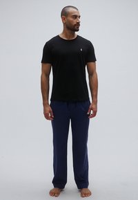 Črna bombažna majica s kratkimi rokavi, kombinirana z mornarsko modrimi bombažnimi hlačami, brez obuvala, na svetlo sivi podlagi.