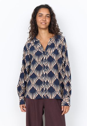 Woman with curly hair wears a patterned button-up blouse with blue and beige geometric shapes and dark brown wide-leg pants.