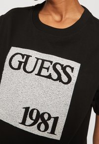 Svart bomull t-shirt med en texturerad, silverfärgad rektangulär logotyp med "GUESS" ovanför och "1981" nedanför i fet svart typografi.