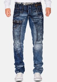 Jeans in denim azzurro chiaro con molteplici tasche, cuciture variate, dettagli strappati e un design slim fit. Sneakers con lacci bianchi in vista.