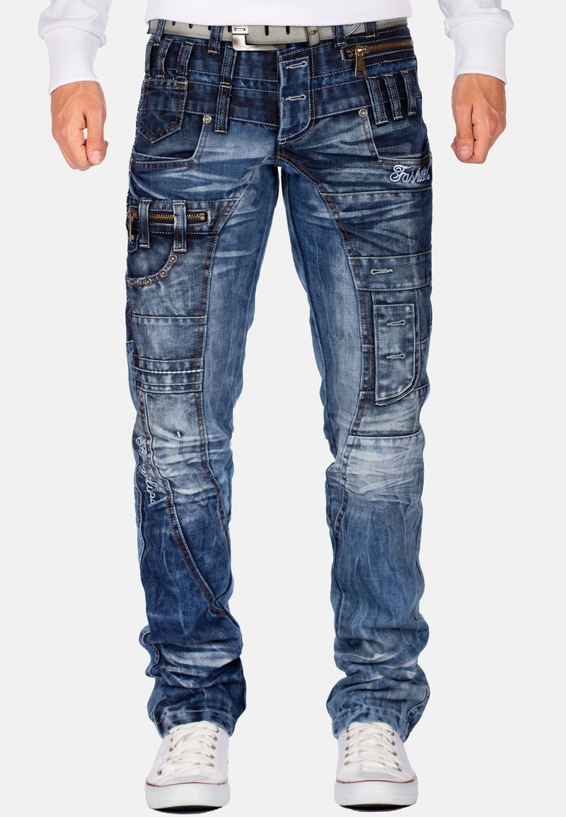 Jeans in denim azzurro chiaro con molteplici tasche, cuciture variate, dettagli strappati e un design slim fit. Sneakers con lacci bianchi in vista.