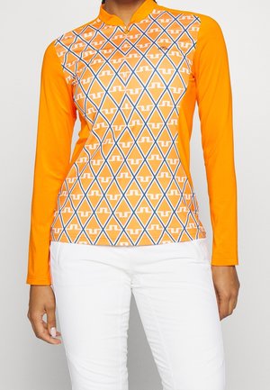 Kobieta ubrana w pomarańczową bluzkę z długim rękawem z geometrycznym wzorem rombów oraz białe spodnie, stojąca na jednolitym tle.