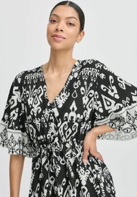 Schwarz-weißen gemustertes Kleid mit kurzen Ärmeln, V-Ausschnitt, Kordelzug an der Taille und Knopfdetails. Der Stoff hat eine strukturierte Oberfläche.