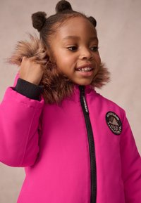 Gaiši rozā pūkains mētelis ar mākslīgās kažokādas kapuci, ar rāvējslēdzēja aizdari un aptuveni apļu logotipa uzšuvumu uz krūtīm. Rievoti aproces.