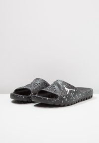 Mustat liukuasut sandaalit, joissa on valkoisia maalaustahroja, tuuletettu päällinen ja valkoinen Jumpman-logo sivuilla, aseteltuna vierekkäin.