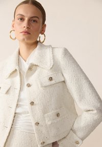 Giacca bianca in tweed con colletto, cinque bottoni decorativi, due tasche sul petto e sottili filamenti nel tessuto.