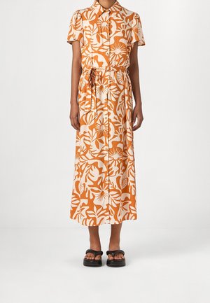 Kvinde iført ankel-lang, kortærmet kjole med knapper foran, med orange og hvid tropisk blomsterprint og matchende stofbælte, sorte sandaler.