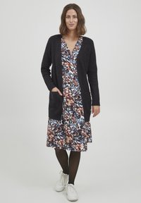Mörkstickad cardigan med ribbat mönster, kombinerad med en blommig klänning i blått, brunt och cream, avslutad med vita sneakers.