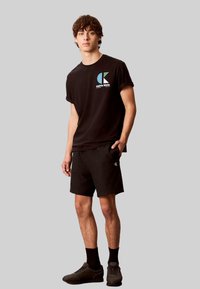Schwarzes Baumwoll-T-Shirt mit farbigem Logo, kombiniert mit schwarzen Shorts. Das Modell trägt schwarze Socken und dunkle Sportschuhe und steht vor einem grauen Hintergrund.