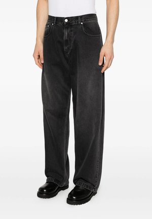 Persona che indossa jeans neri larghi lavati con tasche frontali e scarpe di pelle nere, in piedi davanti a uno sfondo bianco semplice.