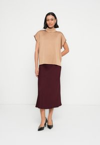 Beige lyhythihainen paita, jossa on niskakiinnitys, yhdistetty viininpunaiseen midihameeseen. Asukokonaisuudessa on sileä pinta ja istuva siluetti.