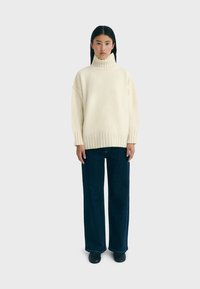Cremefarbener oversized Strickpullover mit hohem Rippenkragen, kombiniert mit weit geschnittenen dunklen Jeans und schwarzen Schuhen. Sanfte Textur, lockere Passform.