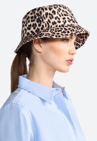 Bucket-Hut mit Leopardenmuster aus weichem Stoff, mit breiter Krempe und schwarzem Logo-Detail. Kombiniert mit einem hellblauen Hemd mit Kragen.