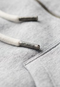 Tessuto felpato grigio chiaro con trama testurizzata; presenta lacci bianchi con occhielli metallici argentati etichettati "SUPERDRY®." Dettaglio in primo piano.