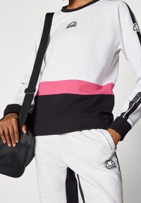 Sudadera gris y negra con una franja horizontal rosa, que presenta un logo en el pecho. Combinada con pantalones grises y una bolsa cruzada negra.
