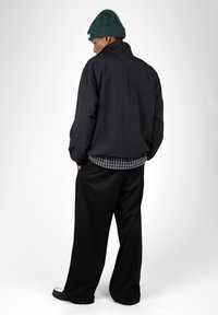 Jeune homme en veste noire et pantalon large noir, les mains dans les poches, portant un bonnet vert tricoté et une chemise à carreaux en dessous.