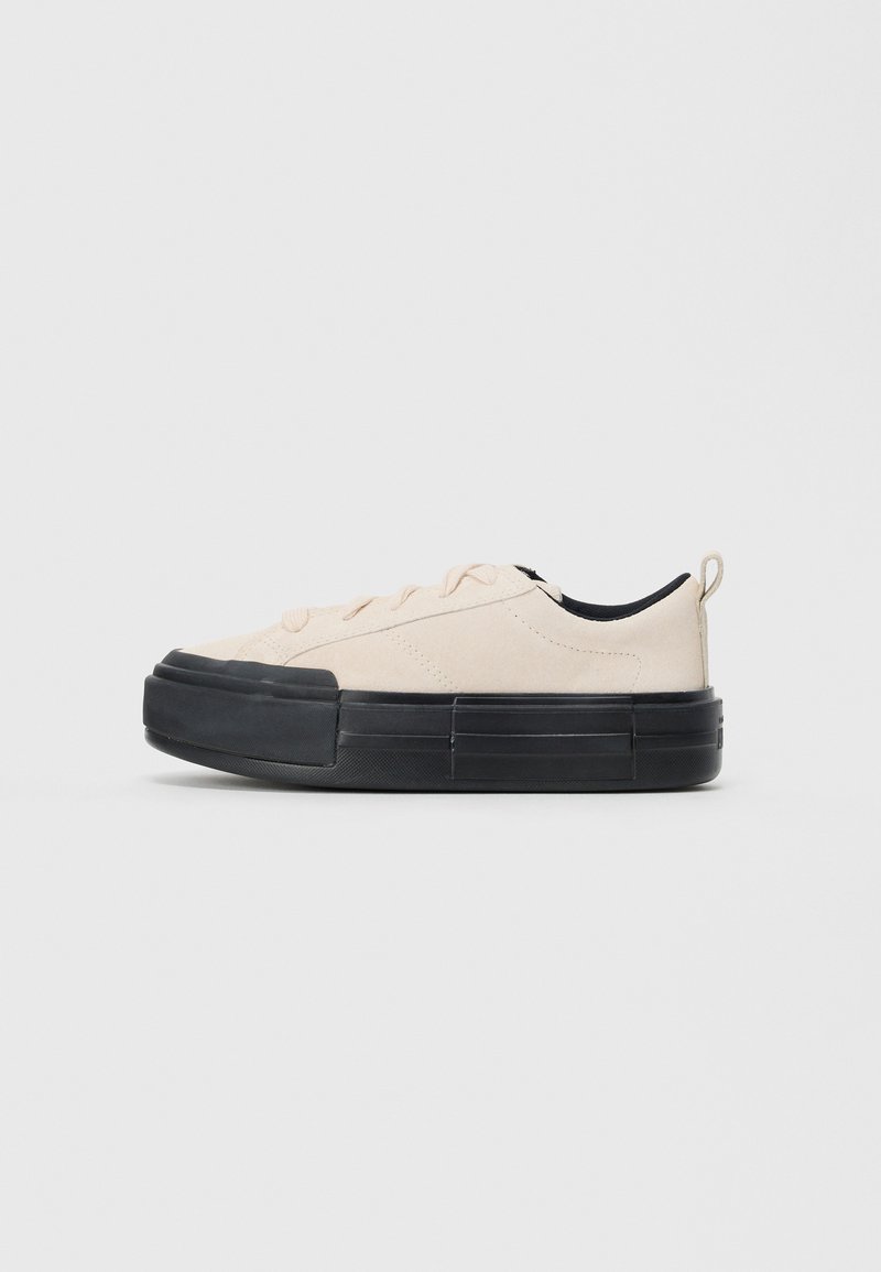 Lave topp beige sneakers med tykk svart såle, snøring foran, stoffpull-tab på hælen og svart innvendig fôr.