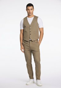 Gilet marrone con una texture raffinata, dotato di cinque bottoni e due tasche frontali, abbinato a pantaloni affusolati dello stesso colore e sneakers bianche.