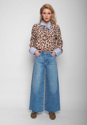 Woman wearing leopard print blouse with blue striped collar and cuffs, wide-leg blue jeans, and brown moccasin shoes, standing with hands in pockets.