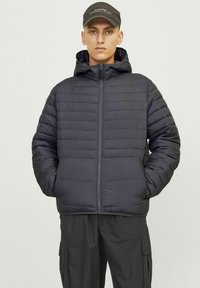 Sort puffjakke med hætte, der har quiltet design, lynlås lukning og sidelommer. Bæres med mørke cargo bukser og en kasket.