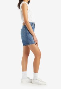 Džínové šortky s vysokým pasem v středně modré barvě, s designem pěti kapes a jemným prošíváním. Kombinovány s bílou sleeveless halenou a bílými teniskami.