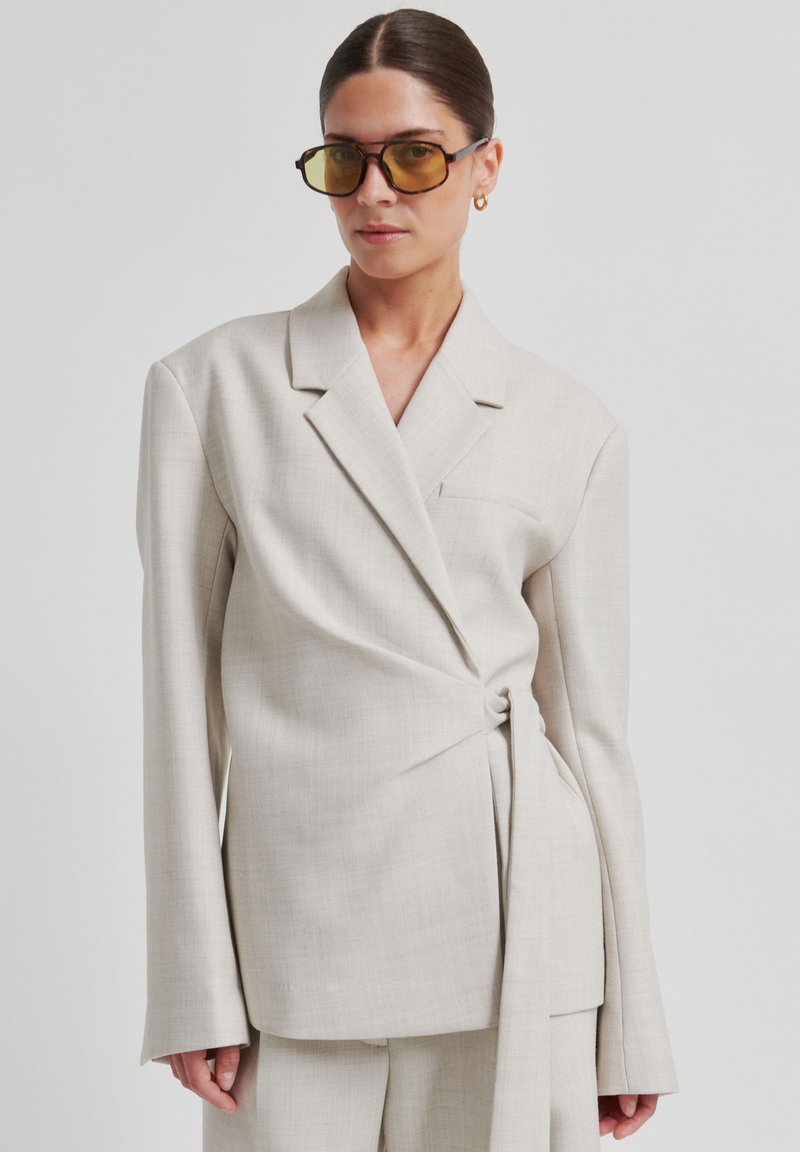 Blazer beige clair cintré avec un design portefeuille, manches larges et un détail de nœud sur le côté. Tissu lisse avec une texture subtile.