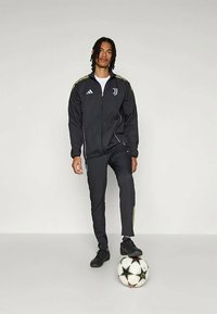 Svart träningsdräkt med gula accenter, zip-up jacka, figurnära byxor och svarta sneakers. En fotboll med svarta stjärnor ligger vid fötterna.