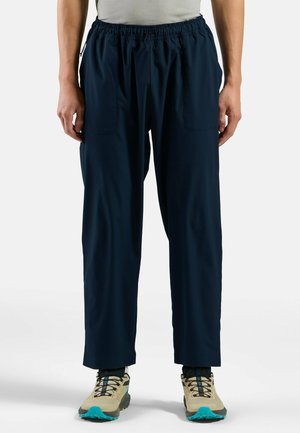 Marineblaue, leichte Hose mit elastischem Bund, zwei seitlichen Taschen und lockerer Passform, kombiniert mit tanfarbenen und türkisfarbenen Wanderschuhen.