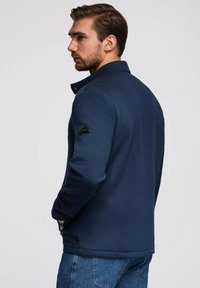 Marinblå texturerad jacka med krage och dragkedja, med en triangulär logotyp på vänster ärm. I kombination med blå jeans, sedd från sidan.