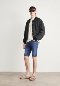 Giubbotto bomber nero, camicia bianca, pantaloni corti in denim blu e sandali beige. Il modello è in piedi su uno sfondo neutro, indossando occhiali da sole rettangolari.