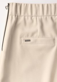 Beige-housut, joissa on teksturoitu kangas, joustava vyötärö, sivuvetoketju ja pieni takatasku, jossa on metallinen logo levytys.