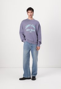 Purple sweatshirt with "TOMMY JEANS NEW YORK CITY" in teal lettering, paired with light blue wide-leg jeans and black shoes.