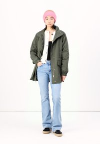 Parka verte avec capuche, chemise blanche à boutons, haut noir, jean flared bleu, bonnet en tricot rose et baskets noires avec semelles beiges.