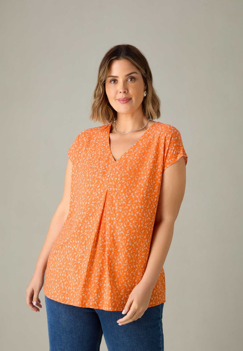 Top floreale arancione con maniche corte e scollatura a V, caratterizzato da una vestibilità ampia e da una leggera arricciatura al centro. Abbinato a jeans blu.