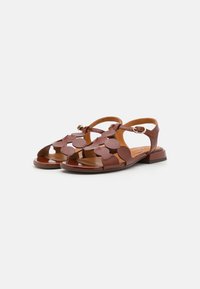 Des sandales en cuir marron présentent un design à bout ouvert avec de grandes découpes en forme de fleurs, un petit talon large et une bride de cheville ajustable.