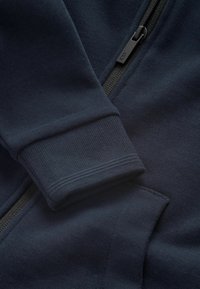 Granatowa bluza z kapturem na zamek błyskawiczny, wykonana z teksturowanej tkaniny, z prążkowanym mankietem i bocznymi kieszeniami. Zawiera czarny zamek z logo jako uchwyt.