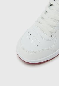 Λευκό αθλητικό παπούτσι με λείο δερμάτινο επάνω μέρος, στρογγυλή μύτη, διάτρητες λεπτομέρειες στο επάνω μέρος και κόκκινη εξωτερική σόλα από καουτσούκ. Οι κορδόνια είναι λευκά.