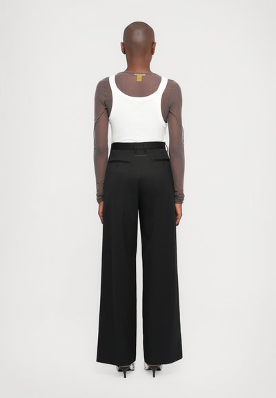White tank top layered beneath a sheer long-sleeve top, black wide-leg pants, and black heels. Notable elements include a tag on the shirt.