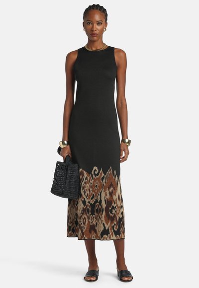 Žena oblečená v čiernych šatách bez rukávov s hnedým a béžovým vzorovaným lemom, drží čiernu tkanú kabelku a má na nohách čierne šľapky.
