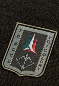 Patch ricamato con sfondo nero, forma a scudo e la scritta "FRECCE TRICOLORI." Include arrow verdi, bianchi e rossi e una grafica di un arco.