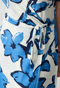 Wikkeljurk met een blauw en marineblauw bloemenpatroon op witte stof, voorzien van een strikceintuur in de taille en subtiele textuurstiksels.