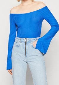 Blue ribbed long-sleeve off-the-shoulder top with bell sleeves, paired with high-waisted light denim jeans featuring a zipper detail.