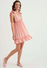 Rosa Kleid mit enganliegendem Oberteil, tiefem V-Ausschnitt und Rüschensaum. Verfügt über Bindebänder und ein gepunktetes Muster. Getragen mit passenden rosa Ballerinas.