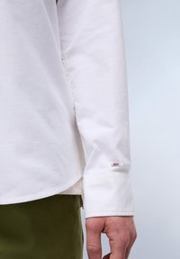 Camicetta bianca a maniche lunghe con orlo a polsino, caratterizzata da un piccolo tag con logo sulla manica. Tessuto di cotone morbido con una vestibilità comoda.