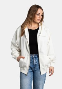 Bílá mikina na zip vyrobená z měkké tkaniny, s volným střihem, velkými předními kapsami a žebrovanými manžetami, v kombinaci s džíny s vysokým pasem.