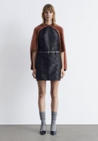 Schwarzes Kleid mit floralem Muster, tailliert mit einem grauen Gürtel. Eine braune Mohair-Cape, graue Socken und schwarze spitze Schuhe vollenden den Look.