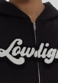 Sweat à capuche noir zippé avec un logo "Lowlight" brodé en blanc, doux et texturé, sur le devant. Dispose d'une capuche et d'une fermeture éclair argentée.