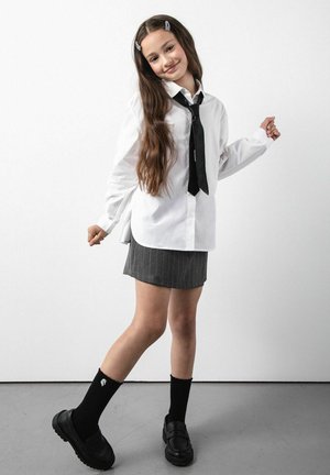 Biała koszula na guziki z długimi rękawami, czarny krawat, szara spódnica w drobną prążkowaną kratę, czarne podkolanówki i czarne mokasyny. Długie, falowane brązowe włosy.