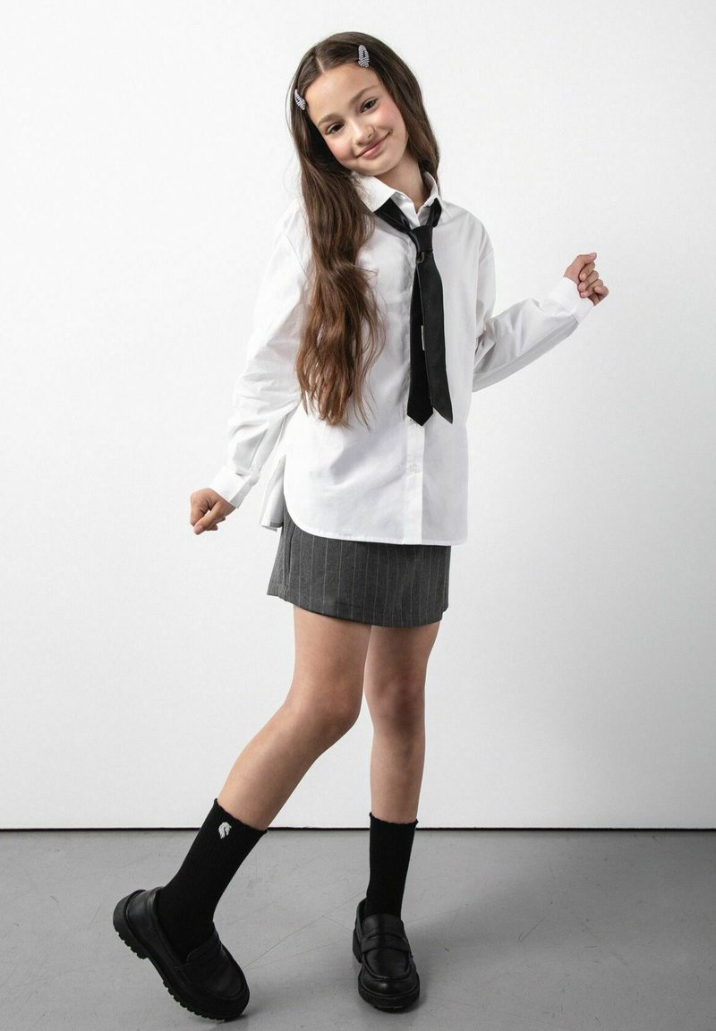 Biała koszula na guziki z długimi rękawami, czarny krawat, szara spódnica w drobną prążkowaną kratę, czarne podkolanówki i czarne mokasyny. Długie, falowane brązowe włosy.